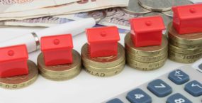 A red houses sitting on a coins, with a pen and calculator to symbolize house finance in the UK