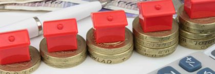 A red houses sitting on a coins, with a pen and calculator to symbolize house finance in the UK